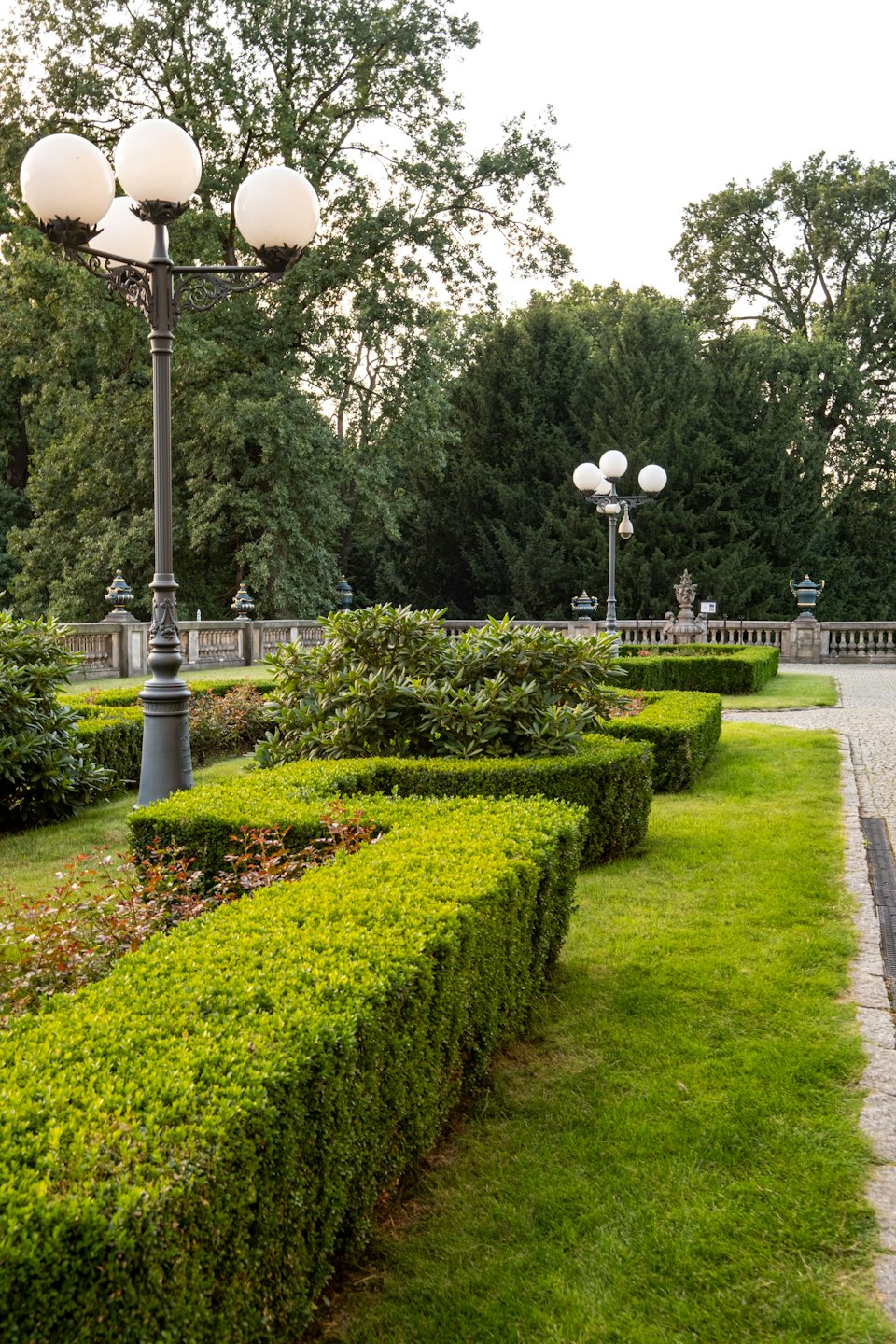 Formeller Garten mit geschnittenen Hecken, Straßenlaternen und Steinbalustrade.